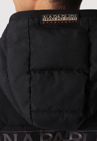 Sort polstret jakke med en struktureret hætte. Har et fremtrædende mærke med "NAPAPIRI" og "geographic" i kontrastfarver. Glat tekstur.