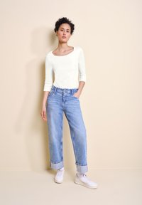 Hvid langærmet top med en rund halsudskæring, parret med lysblå højtaljede jeans med opfoldede manchetter og hvide sneakers. Ensfarvet baggrund.