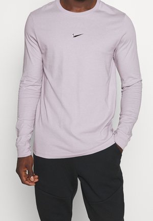 Mężczyzna w jasnofioletowej koszulce Nike z długim rękawem z czarnym logo Swoosh na środku klatki piersiowej i czarnych spodniach, stoi z rękami swobodnie opuszczonymi.