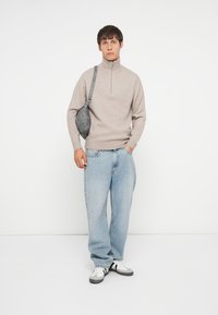 Jersey beige con cremallera, jeans de corte relajado de color azul claro, zapatillas negras y blancas, y un bolso cruzado gris. Diseño simple, textura suave.