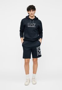 Marinblå set med bomullshoodie och shorts med logotryck framtill. Slät textur, dragsko i huvan och fickor på shortsen.