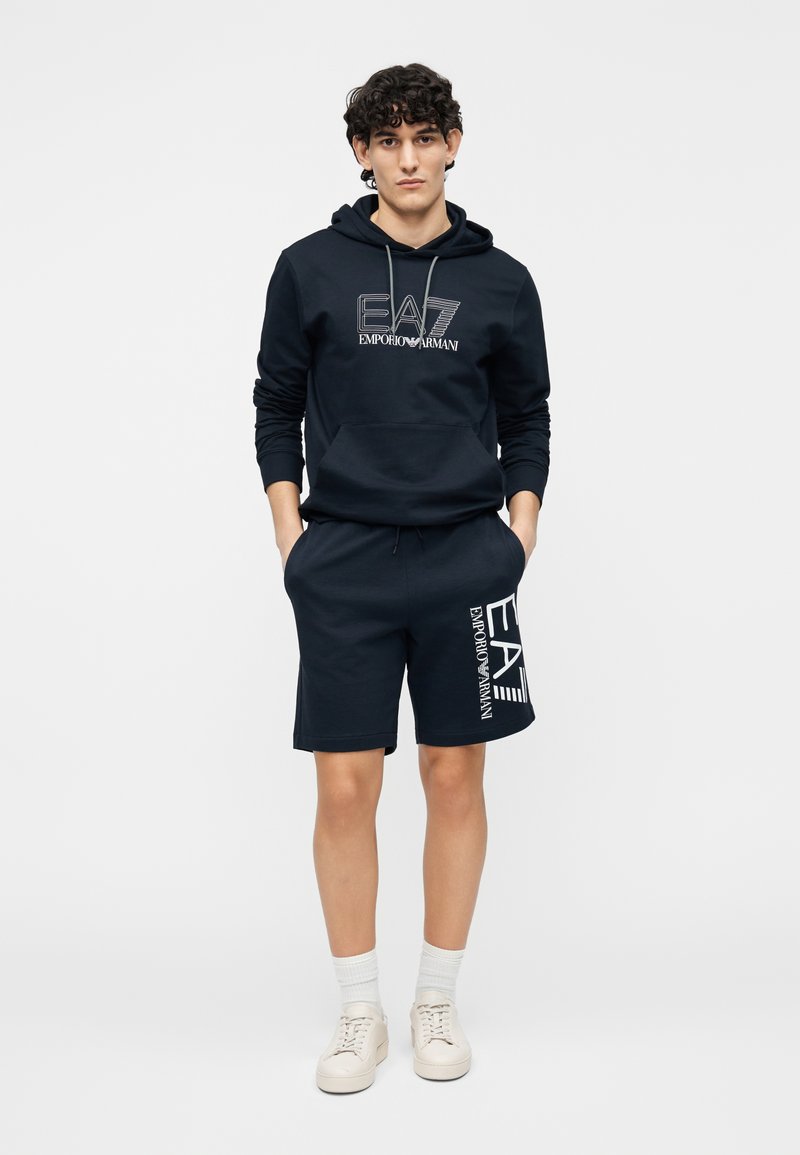 Marinblå set med bomullshoodie och shorts med logotryck framtill. Slät textur, dragsko i huvan och fickor på shortsen.