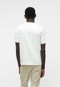 Biela bavlnená tričko s krátkymi rukávmi a okrúhlym výstrihom, s jednoduchou textúrou a rovnými okrajmi, nosené s béžovými nohavicami.