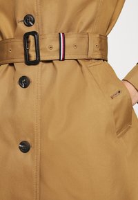 Jasnobrązowy płaszcz trench z paskiem w talii, z czarnymi guzikami i detalami kieszeni. Pasek ma akcent w paski w kolorze czerwonym, białym i niebieskim.