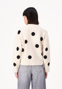 Krämfärgad tröja med stora svarta fluffiga prickar, ribbad mudd och nederkant. Bärs tillsammans med löst sittande ljusgråa jeans. Bakvy visas.