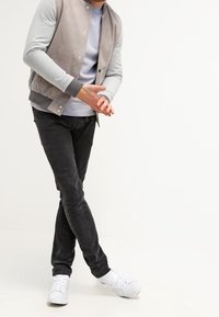 Hombre con chaqueta gris y beige, camisa gris, jeans negros y zapatillas blancas, de pie con las piernas cruzadas y las manos entrelazadas.