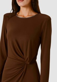 Vestito marrone a maniche lunghe con scollo tondo e dettaglio argentato. Il tessuto è liscio con un dettaglio drappeggiato a torsione in vita.