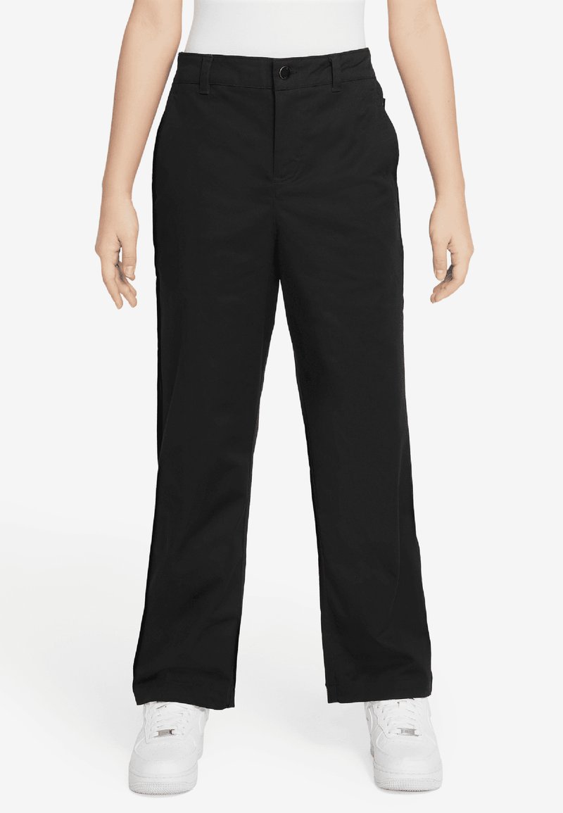 Pantalons noirs à jambe large en tissu léger avec une ceinture, des poches avant et des détails de plis subtils ; associés à des baskets blanches.