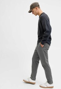 Grauer Pullover mit geripptem Saum und dunkler Struktur, kombiniert mit hellgrauen Hosen und weißen Sneakern mit braunen Sohlen, Model im Profil.