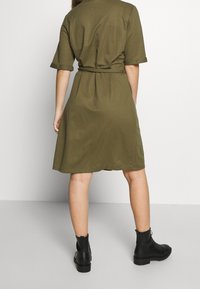 Robe courte à manches courtes de couleur vert olive avec une taille nouée, une jupe évasée et une texture lisse, associée à des bottines noires à semelle épaisse.