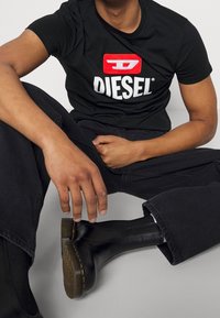 Svart bomullströja med röd och vit Diesel-logotyp, i kombination med svarta denimbyxor och svarta ankelstövlar med ribbat mönster.