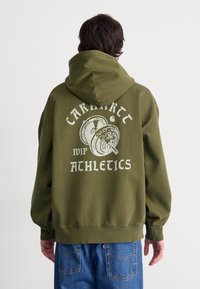 Oliwkowa bluza z kapturem z dużym białym nadrukiem z tyłu, przedstawiającym sztangę oraz tekst "Carhartt VIP Athletics".