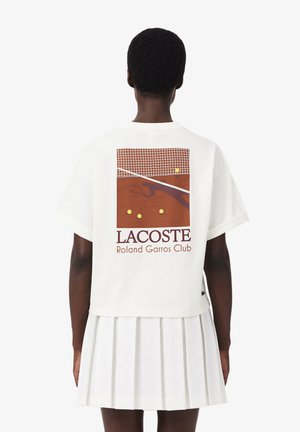 Persona che indossa una t-shirt bianca Lacoste con una grafica del campo da tennis Roland Garros e quattro palline da tennis sul retro, abbinata a una gonna pieghettata bianca.