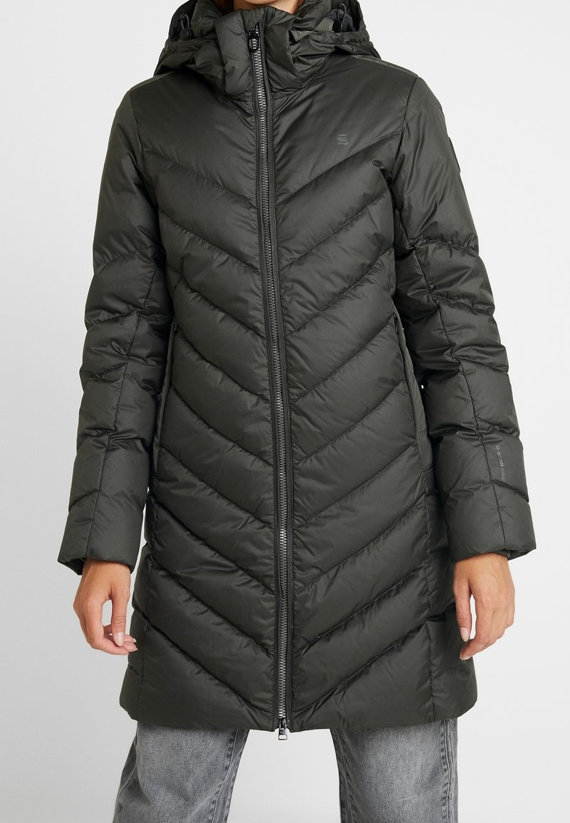 Mörkgrön puffjacka med huva, med ett chevronmönster, dragkedjestängning och långa ärmar. Materialet verkar slätt och isolerat.