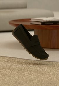 Μαύρο δερμάτινο μοκασίνι με καφέ λαστιχένια σόλα αιωρείται πάνω από μπεζ χαλί, δίπλα σε ξύλινο τραπεζάκι καφέ με στοιβαγμένα βιβλία.