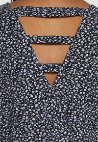 Top en tissu noir avec de petits motifs floraux blancs et bleus ; présente un dos en forme de V avec trois bretelles horizontales pour plus de détail.