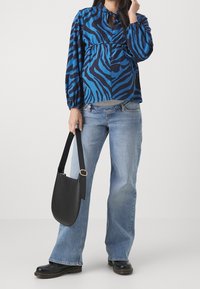 Blouse à rayures zébrées bleues et noires avec un col éclaire, associée à un jean évasé bleu clair et un sac à bandoulière noir. Des chaussures noires complètent la tenue.