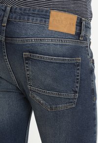 Denimjeans bakvy med en brun Pier One-logotyp, synlig sömnad och en randig skjorta instoppad i midjan.
