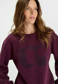 Burgundy sweatshirt with a black oversized logo, long sleeves, and a crew neckline; made from soft cotton blend material.