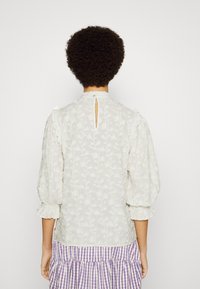Blouse blanche brodée avec un dos en forme de clé, manches bouffantes et accents à volants, associée à une jupe à carreaux violette et blanche.