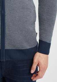 Suéter de cremallera azul marino con un patrón de tejido texturizado de líneas diagonales, puños y dobladillo acanalados. Mano posicionada en el costado, vistiendo jeans oscuros.