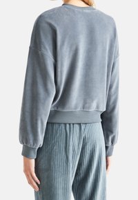 Person som bär en blågrå velourtröja med ribbade manschetter och nederkant, i kombination med matchande strukturerade byxor, sedd bakifrån.