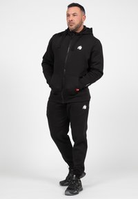 Zwarte rits-hoodie en bijpassende joggers van zacht materiaal. De hoodie heeft een klein wit logo op de borst; de joggers hebben accenten op de zakken.