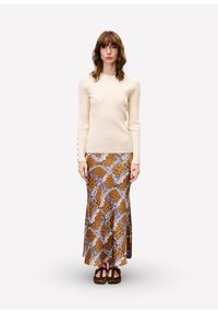 Femme debout portant un pull côtelé crème, une jupe maxi à motifs marron et bleu, et des chaussures plates marron foncé sur un fond clair et uni.