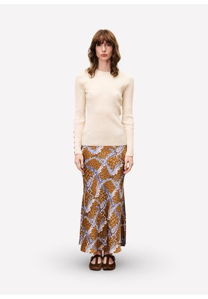 Femme debout portant un pull côtelé crème, une jupe maxi à motifs marron et bleu, et des chaussures plates marron foncé sur un fond clair et uni.