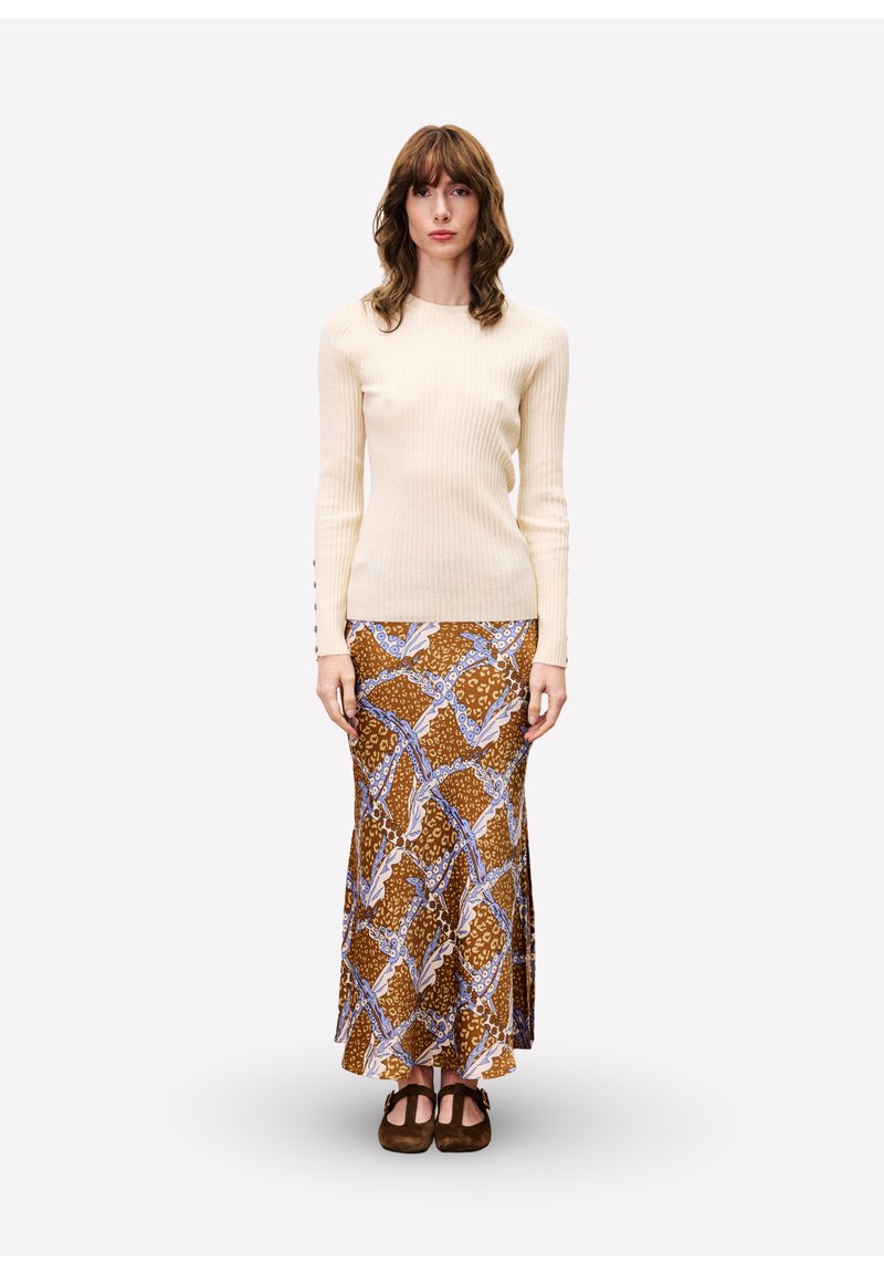 Femme debout portant un pull côtelé crème, une jupe maxi à motifs marron et bleu, et des chaussures plates marron foncé sur un fond clair et uni.