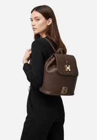Zaino in pelle marrone con texture, chiusura a patta, ferrature dorate e un prominente logo "B" dorato sulla parte anteriore. Spallacci regolabili.