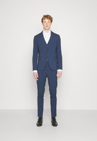 Niebieski garnitur o slim fit, z dwiema guzikami, klapami w kształcie notcha i spodniami z płaskim przodem. Uzupełniony białą koszulą i czarnymi butami.