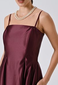 Robe en satin bordeaux avec un corsage ajusté, des bretelles fines et une jupe évasée. Elle présente une texture lisse et un accent en triple rangée de perles.