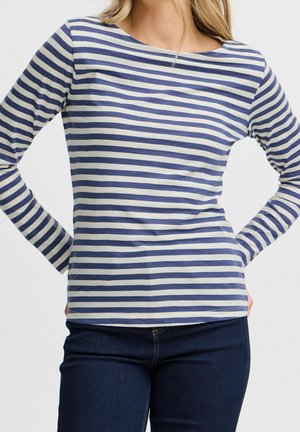 Femme portant un haut à manches longues rayé horizontal bleu et blanc avec un jean bleu foncé, les mains posées sur les hanches sur un fond uni.