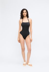 Γυναίκα όρθια, φορώντας μαύρο ολόσωμο μαγιό με ζώνη στο λαιμό και μπεζ σανδάλια τύπου slide, μπροστά από απλό φωτεινό φόντο.