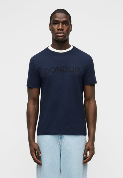 Homme portant un T-shirt Dondup bleu marine à manches courtes sur un sous-vêtement blanc et un jean bleu clair, debout devant un fond uni.
