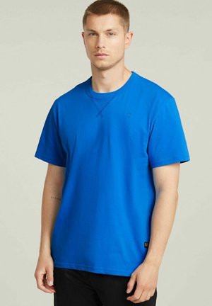 Giovane uomo con capelli castani corti che indossa una maglietta blu a maniche corte senza disegni e pantaloni neri, su uno sfondo neutro.