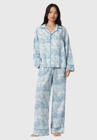 Sinivalkoinen kuviollinen pyjama-asuste, joka on valmistettu pehmeästä kankaasta. Rentomallinen paita, jossa on kaulus, sekä yhteensopivat housut, joissa on joustava vyötärö ja kuminauha lahkeissa.