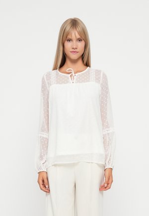 Blouse blanche en tissu à pois transparent, avec un col à cordon, et des accents en dentelle florale sur les manches. Manches longues légèrement bouffantes avec des poignets élastiques.