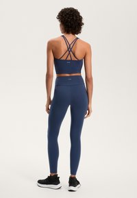 Tengerészeti sportmelltartó keresztbe rakott pántokkal és passzoló leggingsszel, sima, rugalmas anyagból készült. Fekete sportcipővel viselve.