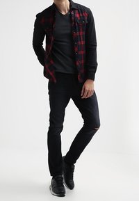 Camisa de cuadros negros y rojos sobre una camiseta negra con cuello en V, combinada con jeans oscuros con un desgarrón en la rodilla. Zapatillas negras completan el conjunto.