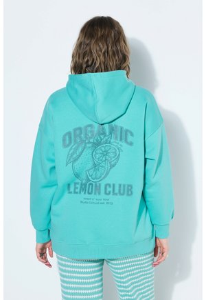 Personne aux cheveux ondulés, de dos, portant un sweat à capuche turquoise avec le texte « Organic Lemon Club » et un graphique de citron, associée à un pantalon à motifs.