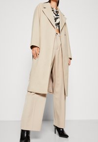 Abrigo beige de mezcla de lana con cuello en muesca, mangas largas y corte recto, combinado con pantalones beige de tiro alto y pierna ancha, y zapatos de tacón negros.