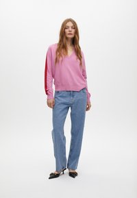 Rosa V-Ausschnitt-Pullover mit roten Akzentstreifen, kombiniert mit hellblauen Denim-Jeans mit Perlenverzierung und schwarzen spitzen Absätzen.