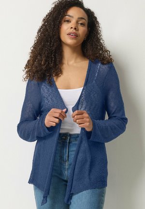 Kvinde med krøllet hår iført en gennemsigtig blå cardigan over en hvid top og blå jeans, som justerer cardiganens forkanter.