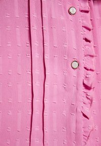 Blusa rosa caratterizzata da un tessuto testurizzato con piccoli punti in rilievo, una patta con bottoni bianchi tondi e un accento arricciato lungo un lato.