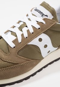 Hnědé semišové a textilní tenisky s bílými tkaničkami, které mají bílou logo panel a texturované detaily. Guma podrážka s černou trakční vrstvou.