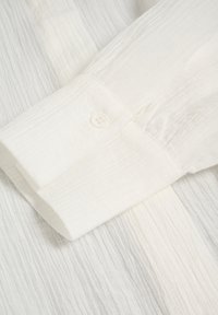 Lähellä oleva valkoinen teksturoitu kangas, jossa näkyy pitkä hiha nappi ja yksittäinen valkoinen nappi kiinnitettynä.