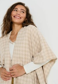 Châle beige à motifs avec une finition texturée. Coupe ample, présentant un design large avec des côtés ouverts et un bord festonné le long de l'ourlet.