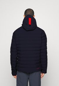 Marinblå pufferjacka med horisontell quiltning, en röd detalj på huvan och en logotags på baksidan. Slät textur och kroppsnära design.